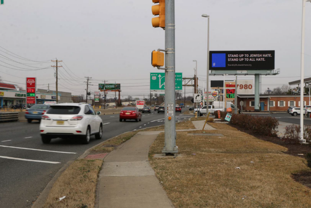 A digital roadside billboard displays an anti-hate message reading, "STAND UP TO JEWISH HATE. STAND UP TO ALL HATE." The billboard has a blue square logo on the left, with the website "StandUpToJewishHate.org" written below. It is mounted on a metal structure above a gas station, along a busy road with cars and traffic signs.