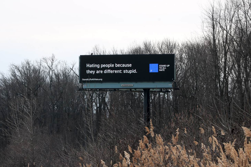 A roadside digital billboard displays an anti-hate message in front of a backdrop of leafless trees. The billboard reads: "Hating people because they are different: stupid." To the right of the text, there is a blue square with the words "STAND UP TO ALL HATE" written next to a hashtag symbol. Below the main text, a website URL "StandUpToAllHate.org" is visible.