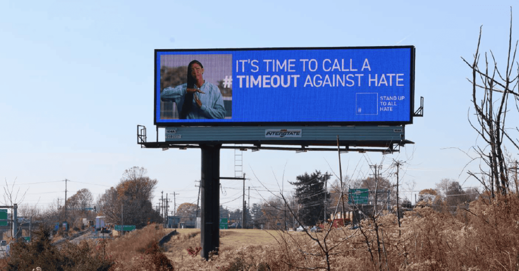 A roadside digital billboard on a tall metal pole displays the message "IT'S TIME TO CALL A #TIMEOUT AGAINST HATE" with a blue background. WNBA star Candace Parker on the left side is making a timeout gesture. The "STAND UP TO ALL HATE" logo is on the right.