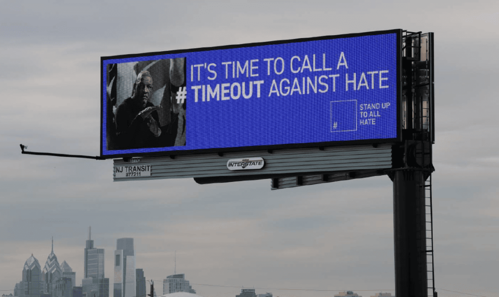 A digital highway billboard with a city skyline in the background displays a message reading, "IT'S TIME TO CALL A #TIMEOUT AGAINST HATE." The billboard has a blue background and features an image of NBA coach Doc Rivers making a timeout gesture. The "STAND UP TO ALL HATE" logo is visible on the right side.