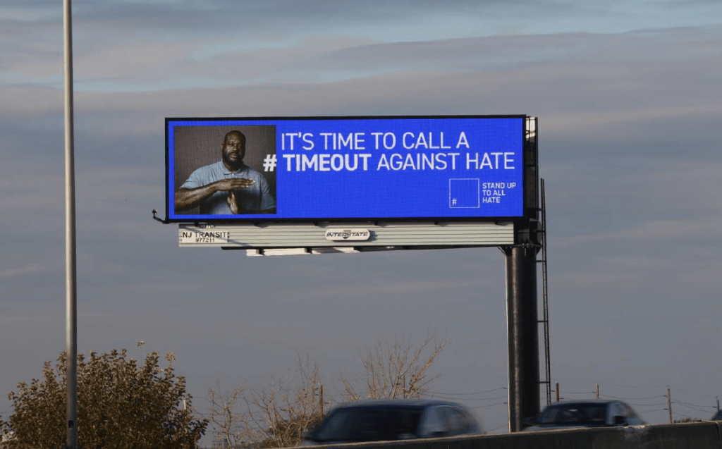 A digital billboard above a highway reads "IT'S TIME TO CALL A #TIMEOUT AGAINST HATE" on a blue background. On the left, an image of NBA legend Shaq O'Neill making a timeout gesture is visible. The "STAND UP TO ALL HATE" logo appears on the right.