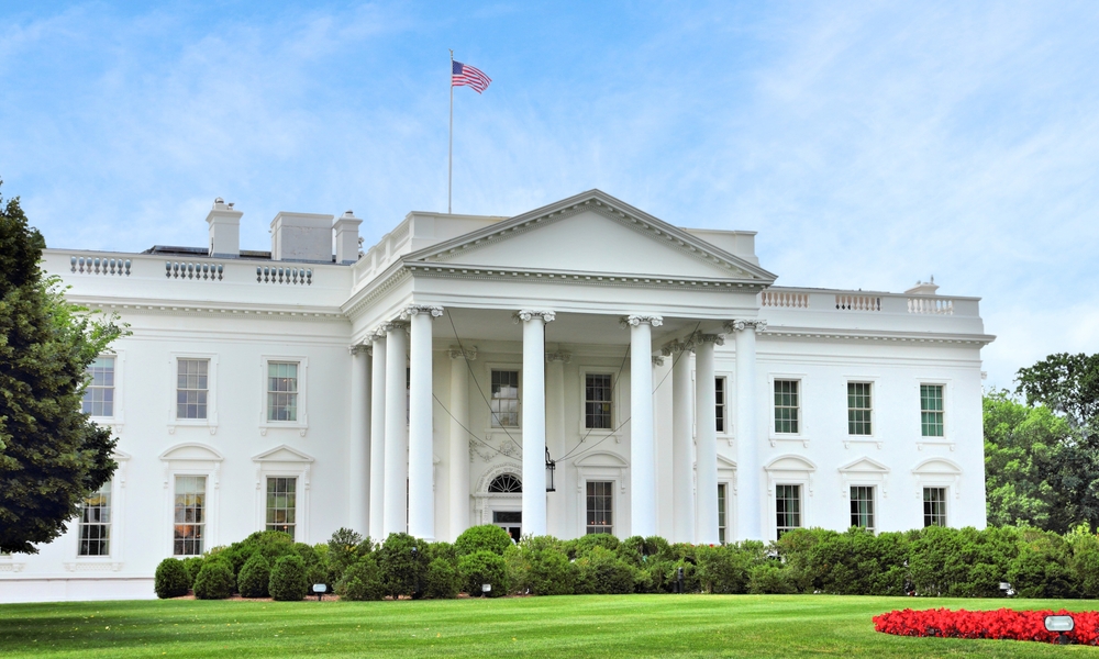 A bright, front-facing view of the White House from the North Lawn, featuring white columns, an American flag atop the building, and manicured greenery in the foreground under a clear blue sky.