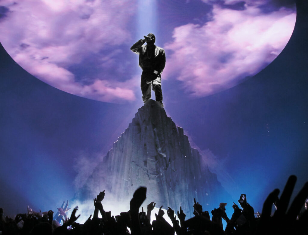Kanye West performs atop a towering, mountain-like stage structure, silhouetted by a bright spotlight and a giant circular backdrop filled with purple clouds. Fans raise their hands and phones in the foreground as dramatic blue and violet lighting fills the arena.