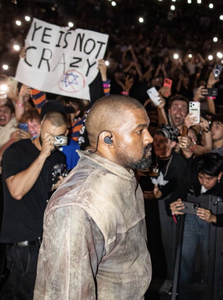 Image from Kanye West concert of Kanye West with a fan in the background holding a sign reading "Ye is not crazy" alongside a Star of David inside a prohibition symbol.