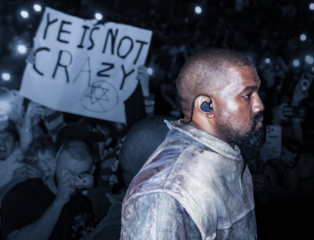 Side-profile view of Ye (formerly Kanye West) on stage wearing an in-ear monitor and a distressed gray jacket. Behind him, a crowd holds up phones and a large handwritten sign that reads, “YE IS NOT CRAZY,” with a Star of David drawn beneath the text. The image has a dark, blue-toned, high-contrast look, with the sign prominently visible in the background.