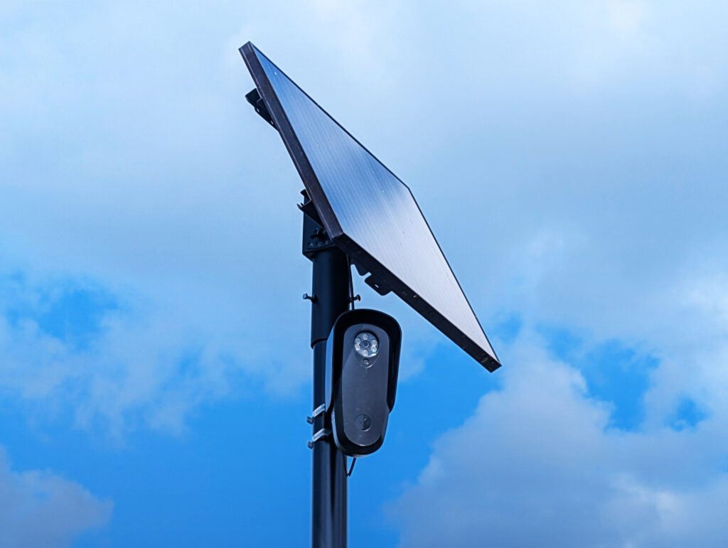 Close-up photo of a pole-mounted surveillance camera beneath a large solar panel, viewed from below against a bright blue, partly cloudy sky.